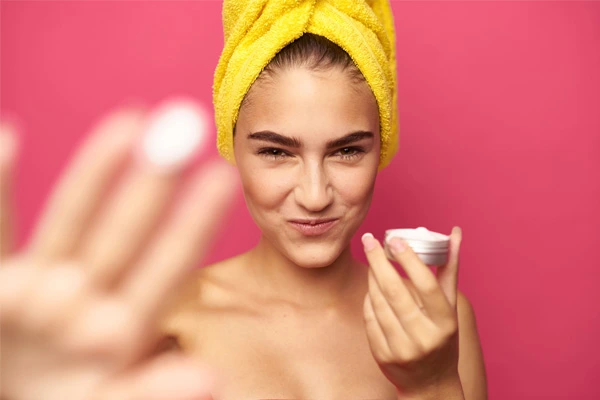 Rozbawiona, młoda dziewczyna z ręcznikiem na głowie trzyma w dłoni krem z peptydami i ceramidami